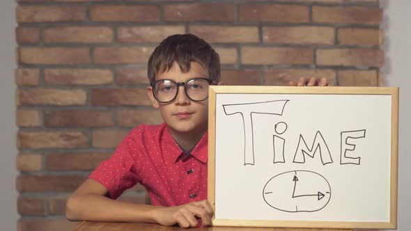 Child Sitting at the Desk Holding Flipchart with Lettering Time on the Background Red Brick Wall