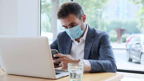 Businessman wearing protective mask using smartphone at laptop in cafe