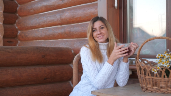 Brunette Woman Sitting At A Table Drinking Tea, On The Veranda Of A Wooden House