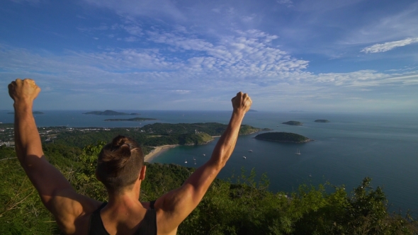 Athletic Young Man Stands with His Hands Up on the Edge of the Mountain