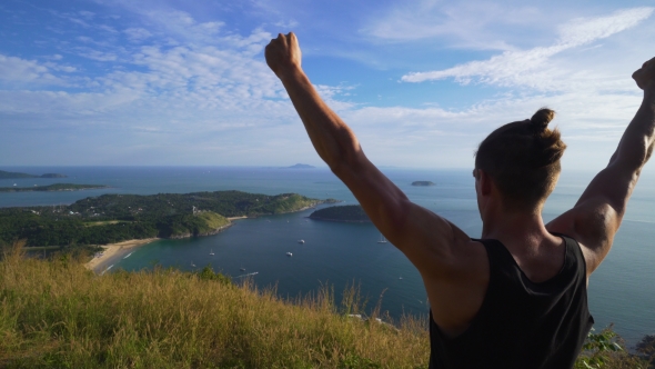 Athletic Young Man Stands with His Hands Up on the Edge of the Mountain