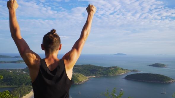 Athletic Young Man Stands with His Hands Up on the Edge of the Mountain