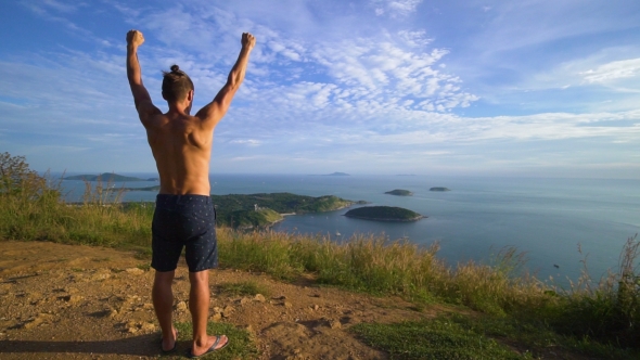 Athletic Young Man Stands with His Hands Up on the Edge of the Mountain