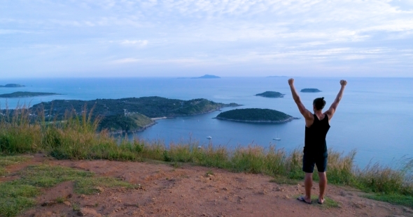 Athletic Young Man Stands with His Hands Up on the Edge of the Mountain