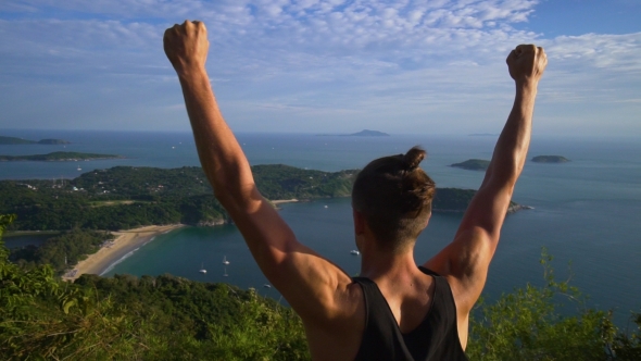 Athletic Young Man Stands with His Hands Up on the Edge of the Mountain.