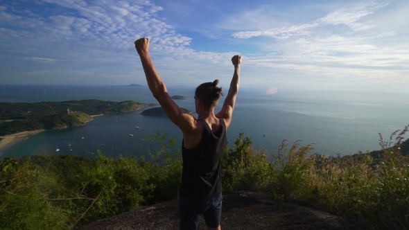 Athletic Young Man Stands with His Hands Up on the Edge of the Mountain.