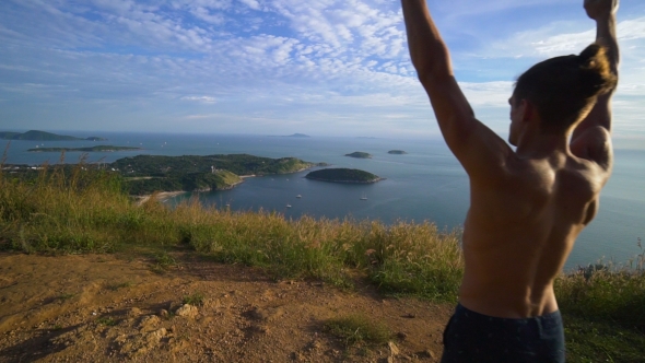 Athletic Young Man Stands with His Hands Up on the Edge of the Mountain.