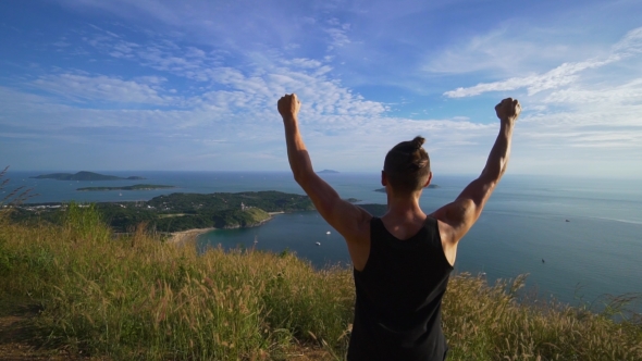 Athletic Young Man Stands with His Hands Up on the Edge of the Mountain.