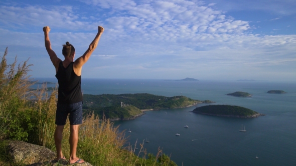 Athletic Young Man Stands with His Hands Up on the Edge of the Mountain.