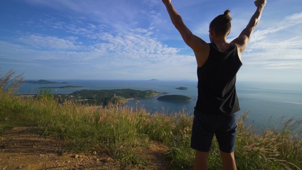 Athletic Young Man Stands with His Hands Up on the Edge of the Mountain.