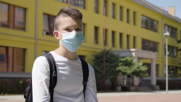 A Caucasian Teenage Boy in a Face Mask Looks at the Camera  a School in the Background