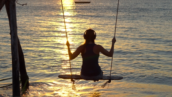 Young Woman in a Wireless Headphones Swings on a Swing Standing in a Sea