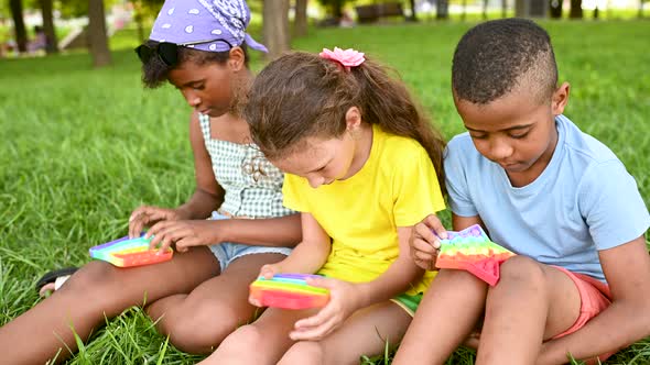 Children of Different Races Sit on the Grass and Play with an Antistress Toy Popit