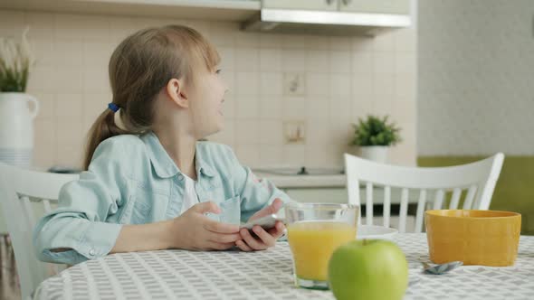 Young Woman Bringing Fresh Croissants To Happy Child Who Is Using Smartphone in Kitchen