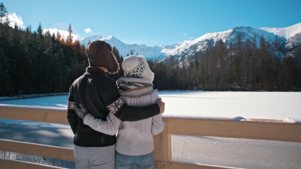 Young Couple Is Standing Near Mountain Lake in Winter Admiring Landskape