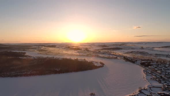 Aerial Over the Frozen Winding River Surrounded with Forest and with a Little Village on a Side