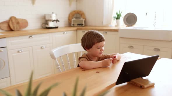 a Little Caucasian Boy Eats Krunchy and Watches Tablet PC at Kitchen Table