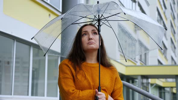 Sad Woman Stands Under a Transparent Umbrella in the Rain on the Background of an Apartment Building