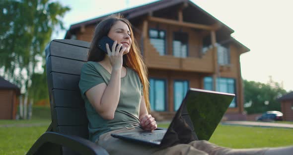 Young Girl Talking on Mobile Phone Sitting in Courtyard of Country House with Laptop