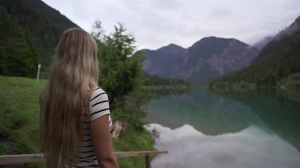 A girl with nice long hair stands in front of a beautiful lake. She is enjoying the scenery with mou