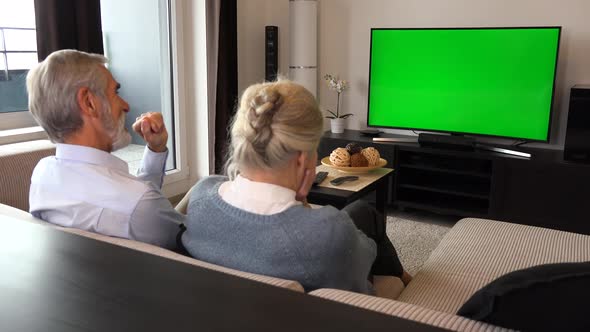 An Elderly Couple Sits on A Couch in A Living Room, Watches Tv with A Green Screen and Laughs