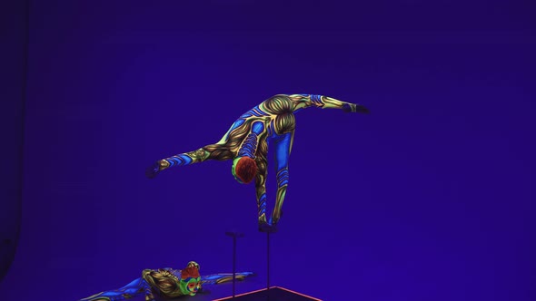 Guy Performs A Stand On One Arm And Does The Splits. Colorful Make Up. Over Blue Background