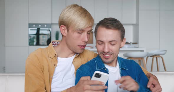 Two Men Homosexual Couple Sit at Home Together in an Embrace on Their Sofa and Using Smartphone