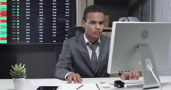 Young African Man in a Modern Office, Working at the Computer, Screens with Financial Infographics