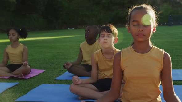 Diverse group of schoolchildren sitting on mats meditating during yoga lesson outdoors