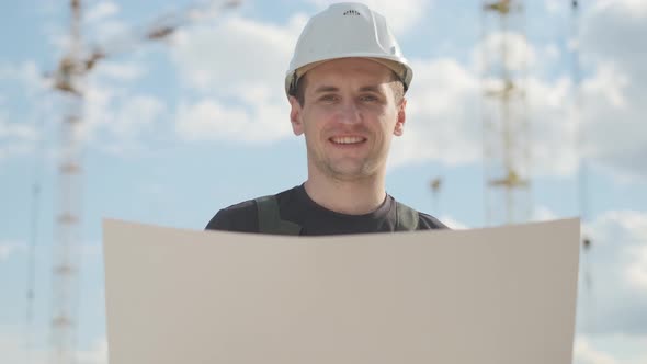 A Man in a Helmet Is Holding a Construction Plan in His Hands and Smiling. A Builder Is Checking