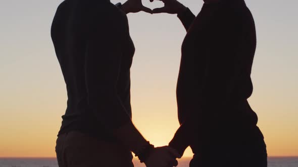 Happy caucasian gay male couple holding hands and making heart shape at sunset on the beach