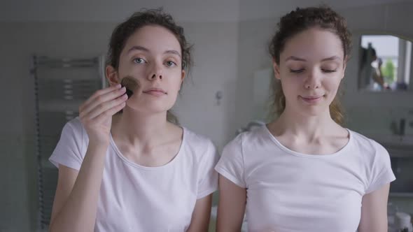 Beautiful Confident Young Slim Woman Applying Face Powder As Twin Sister Putting Cucumber on Skin