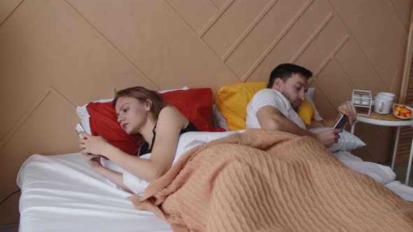 A Married Couple is Lying in Bed with Their Faces Turned Away From Each Other Using Phones