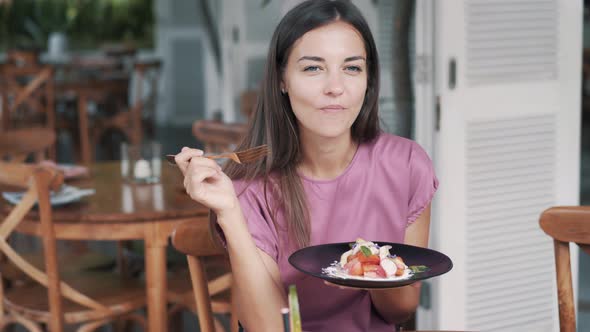 Woman Holds Plate with Vegetable Salad and Eats in Cafe, Looking at Camera