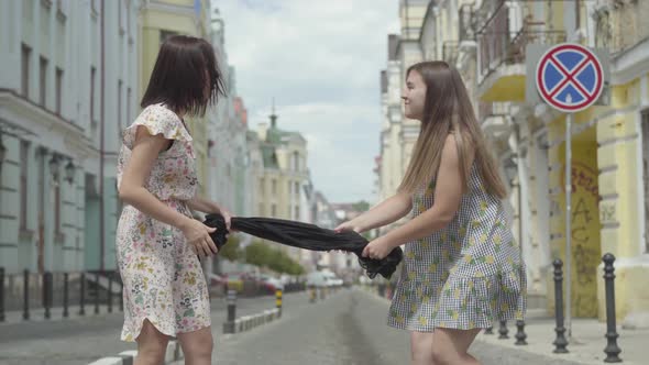 Two Fashionable Girls Holding the Black Dress and Pulling It To Their Sides on the Street