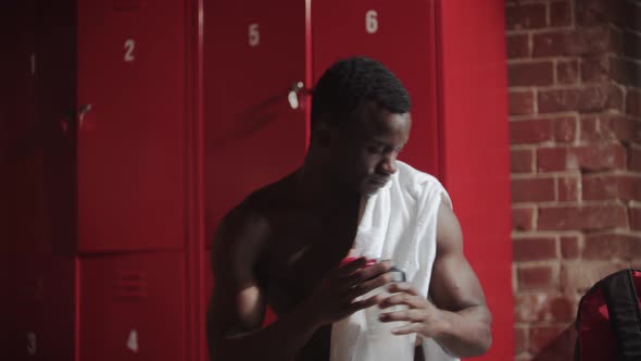 Africanamerican Young Man Drinking Water in the Locker Room and Using His Phone