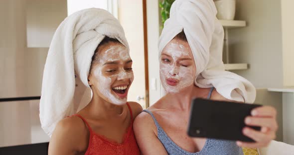Diverse happy female friends wearing towels on heads and cleansing masks taking selfie at home