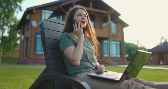 Young Girl Sitting in Lawn Chair at Backyard of Country House with Laptop and Talking on Phone