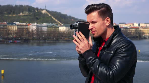 A Young Handsome Man Shoots a Video with a Camera - Closeup - a River and a Quaint Townscape