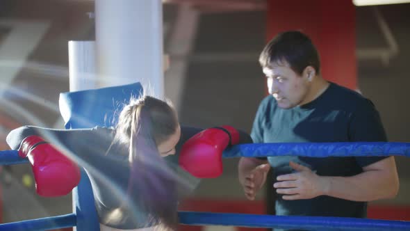 An Attractive Young Woman with Long Hair Resting on the Boxing Ring and Her Trainer Talks To Her