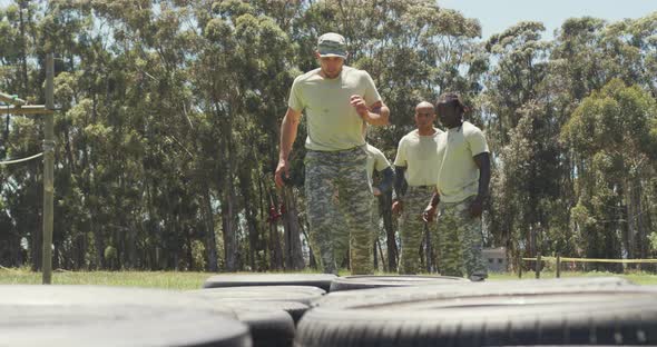 Diverse group of soldiers watching male instructor run through tyres on obstacle course in the sun