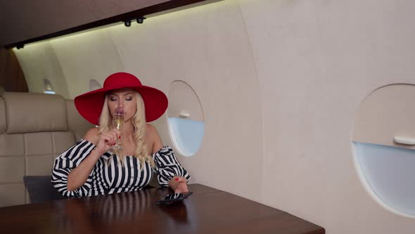 Luxury Blonde Woman with Red Hat is Drinking Champagne During Flight in Her Private Plane