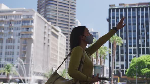 African american woman wearing face mask holding coffee raising hand for a taxi