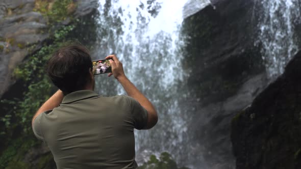 Travel Man with Mobile Phone Shooting Video of Waterfall in Dark Rainforest