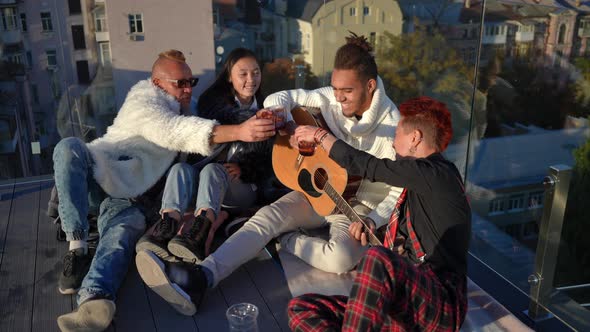 Joyful Adult Friends Clinking Glasses Smiling Listening to Guitar Playing Sitting on Roof in City