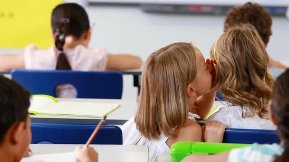 Schoolgirl whispering into her friend s ear in classroom