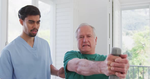 Biracial male therapist talking to senior man exercising with dumbbells at retirement home