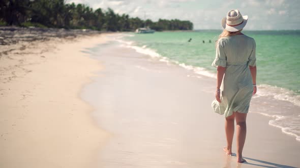 Woman In Hat Walks Along Beach On Caribbean Coast. Woman Relaxing On Seychelles Flowing Dress.