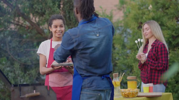 Teenage African American Girl Holding Plate with Raw Meat As Man Putting Burger Patties in Barbecue