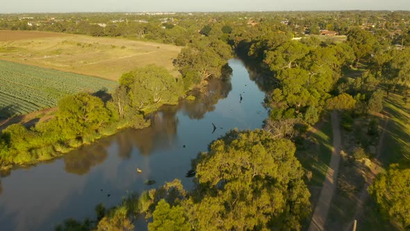 Aerial tracking above natural river between agricultural land and suburban landscape.